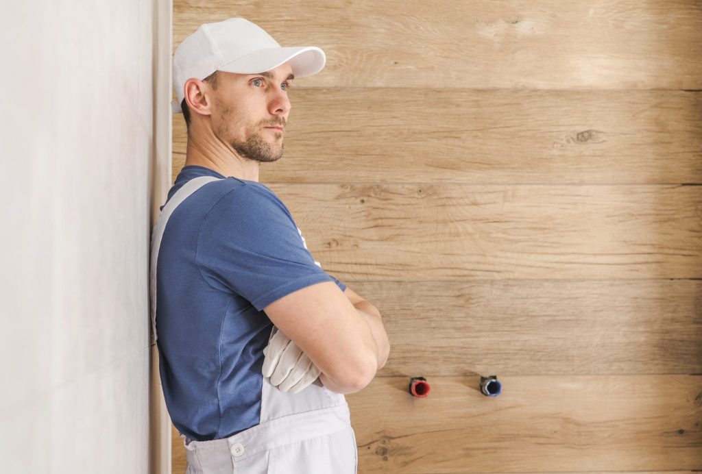 A man in work attire stands with his arms crossed against a wooden wall, looking thoughtfully to the side. He appears to be a home improvement professional.