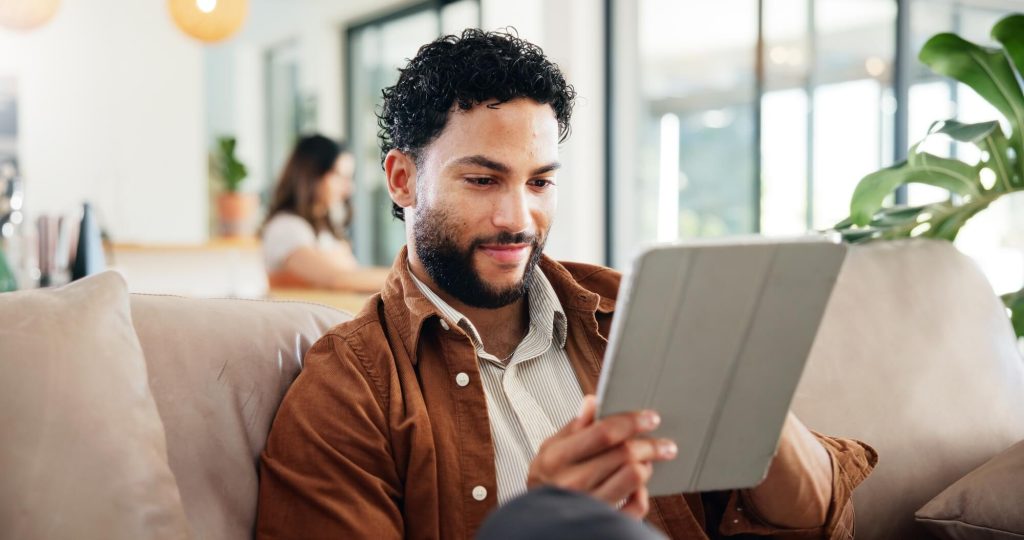 A man sits comfortably on a couch, looking at a tablet with a focused expression. In the background, a woman is seen engaging in another activity.