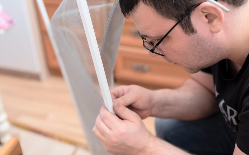 A man is carefully assembling a screen or frame, focusing intently on his work. The setting appears to be a home environment with wooden furniture in the background.